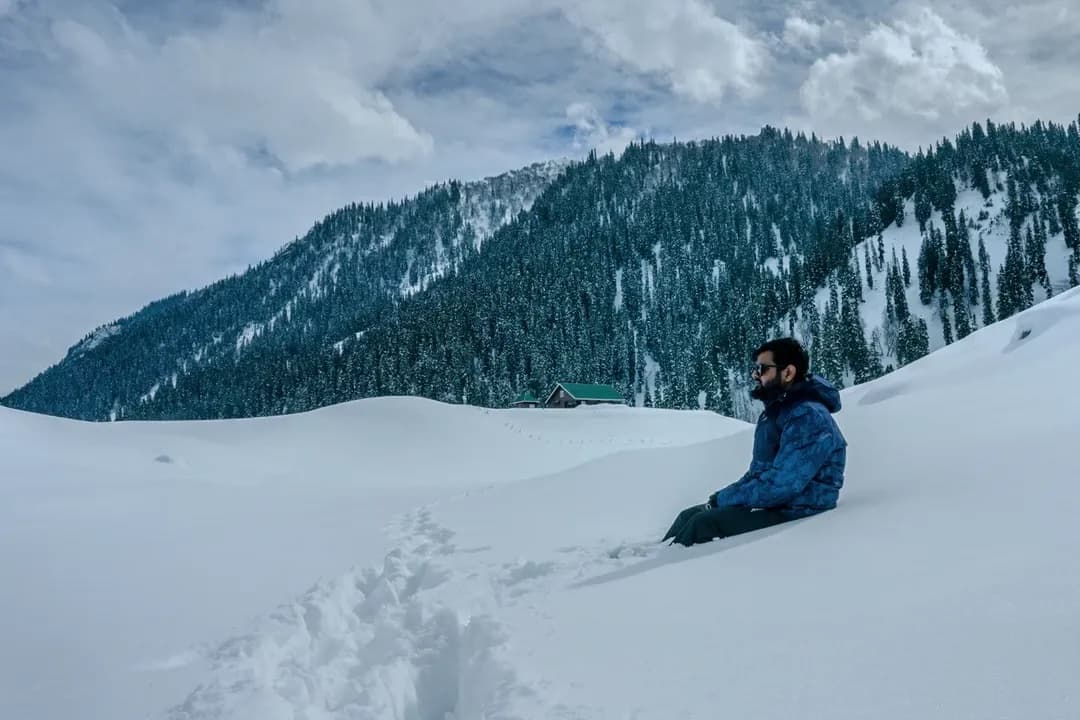 A filmmaker resting in deep snow beneath a pine-covered mountain